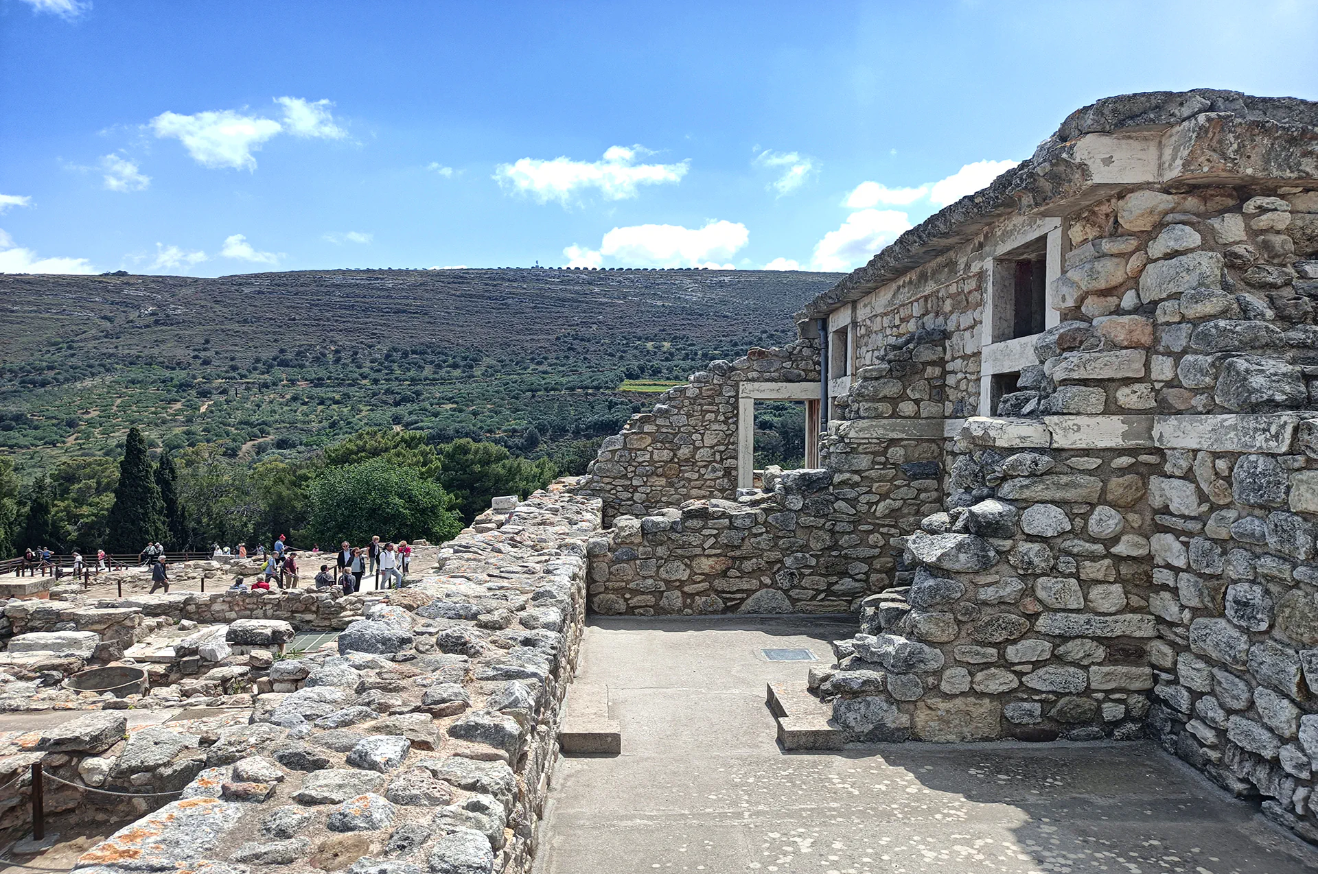 Partially restored stone building at Knossos with tourists and hills in the background.